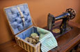 An open woven sewing box with blue padded lining; a green and white checked piece of cloth is lying on the box.