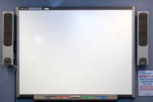 An interactive whiteboard with a speaker on each side. A shelf below the board has a black, red, green and blue marker pen with a whiteboard eraser.