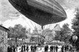 This old hot air balloon has two people riding in the basket; the people watching have clothes from a long time ago.