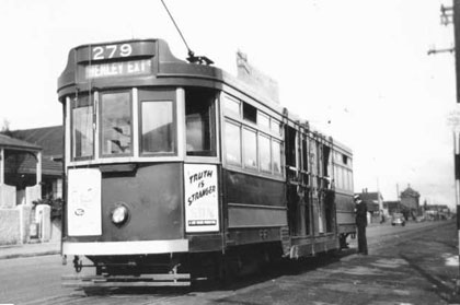 An old electric tram travelling along rails with a conductor standing on the step