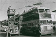 A double decker bus. There are workmen on an old cherry picker fixing the electric wires