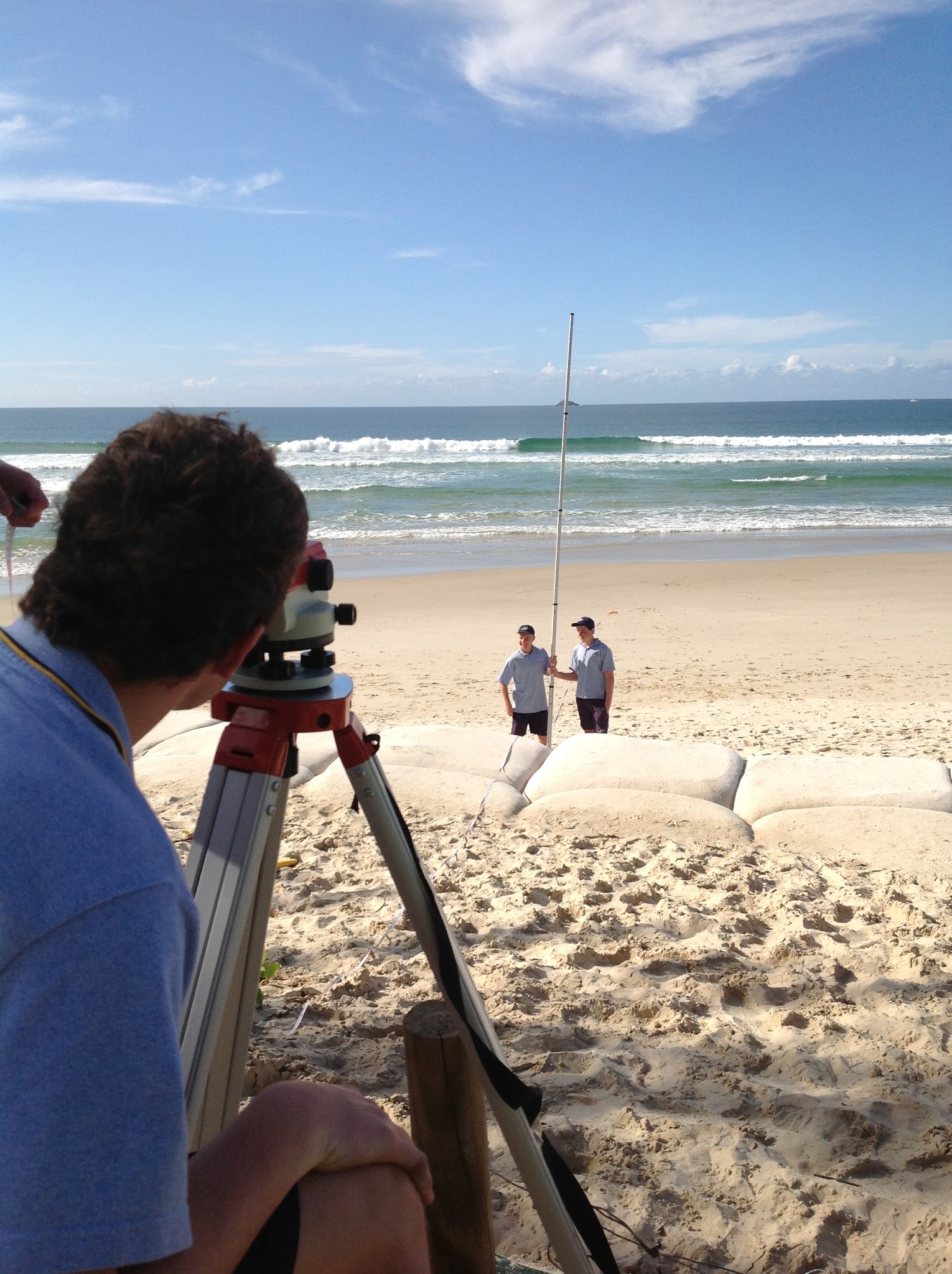 students stand on beach holding a measuring stick whilst another stduent looks at them through surveying equipment. there are sand bags on the beach creating a higher shelf of sand back from the waters edge.