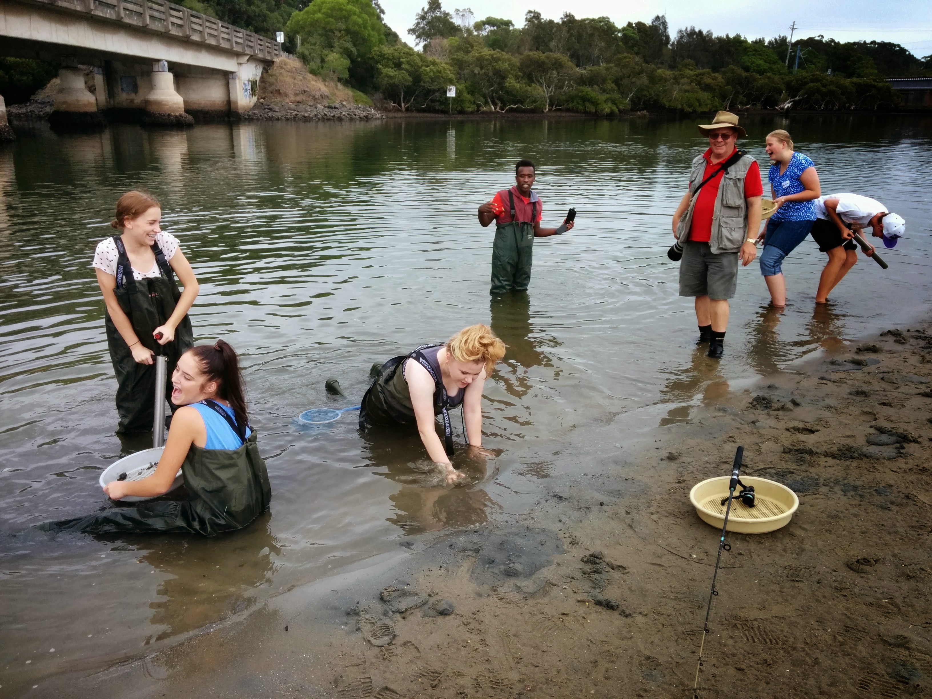 A group of older students wearing protective wading gear are working in the muddy water, they are laughing and having fun.