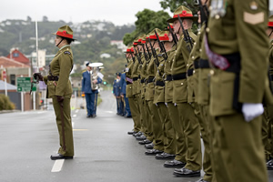 A row of men in military stand at attention; one soldier stands in front with a sabre held vertically in front.