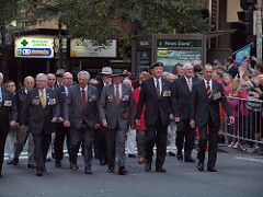 A group of men wearing medals march along a street as people watch from behind barricades.