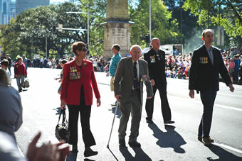 An elderly man walks with the aid of a walking stick down a Sydney street lined with people; he is flanked by a woman on one side and two men on the other; they are all wearing medals An elderly man walks with the aid of a walking stick down a Sydney street lined with people; he is flanked by a woman on one side and two men on the other; they are all wearing medals