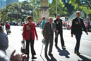 An elderly man walks with the aid of a walking stick down a Sydney street lined with people; he is flanked by a woman on one side and two men on the other; they are all wearing medals An elderly man walks with the aid of a walking stick down a Sydney street lined with people; he is flanked by a woman on one side and two men on the other; they are all wearing medals