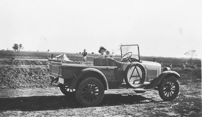 A woman holding a baby in the front seat of a vintage truck in front of the a wide open paddock. A man is standing in the wide open paddock behind them. A woman holding a baby in the front seat of a vintage truck in front of the a wide open paddock. A man is standing in the wide open paddock behind them.